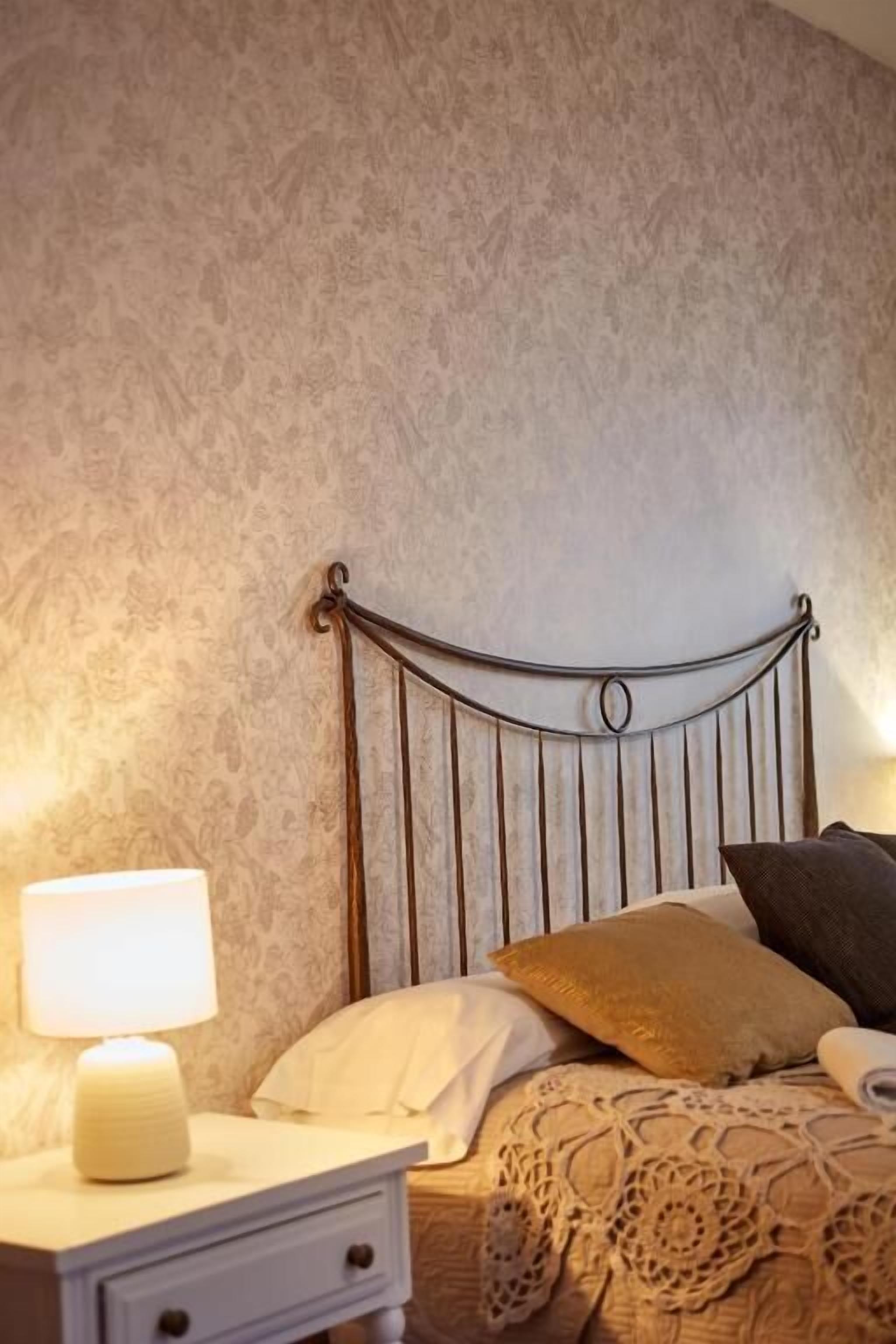 A neatly made bed with a metal headboard, a bedside table with a lamp, and a patterned wall.