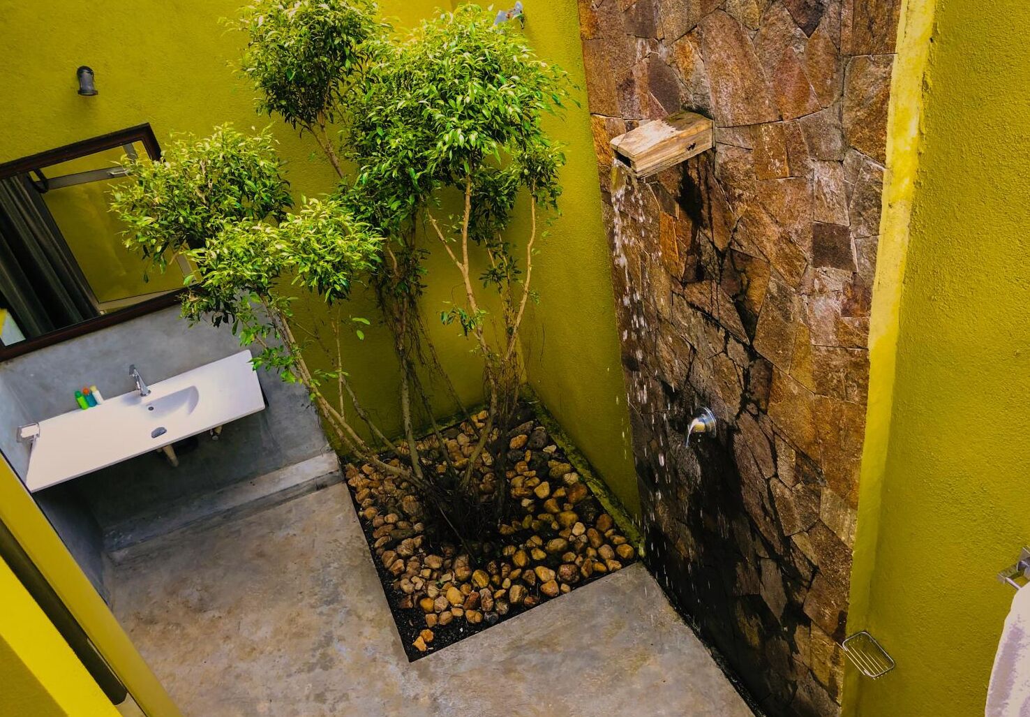 Une salle de bain avec un lavabo blanc, un miroir et une petite cascade décorative adossée à un mur en pierre.