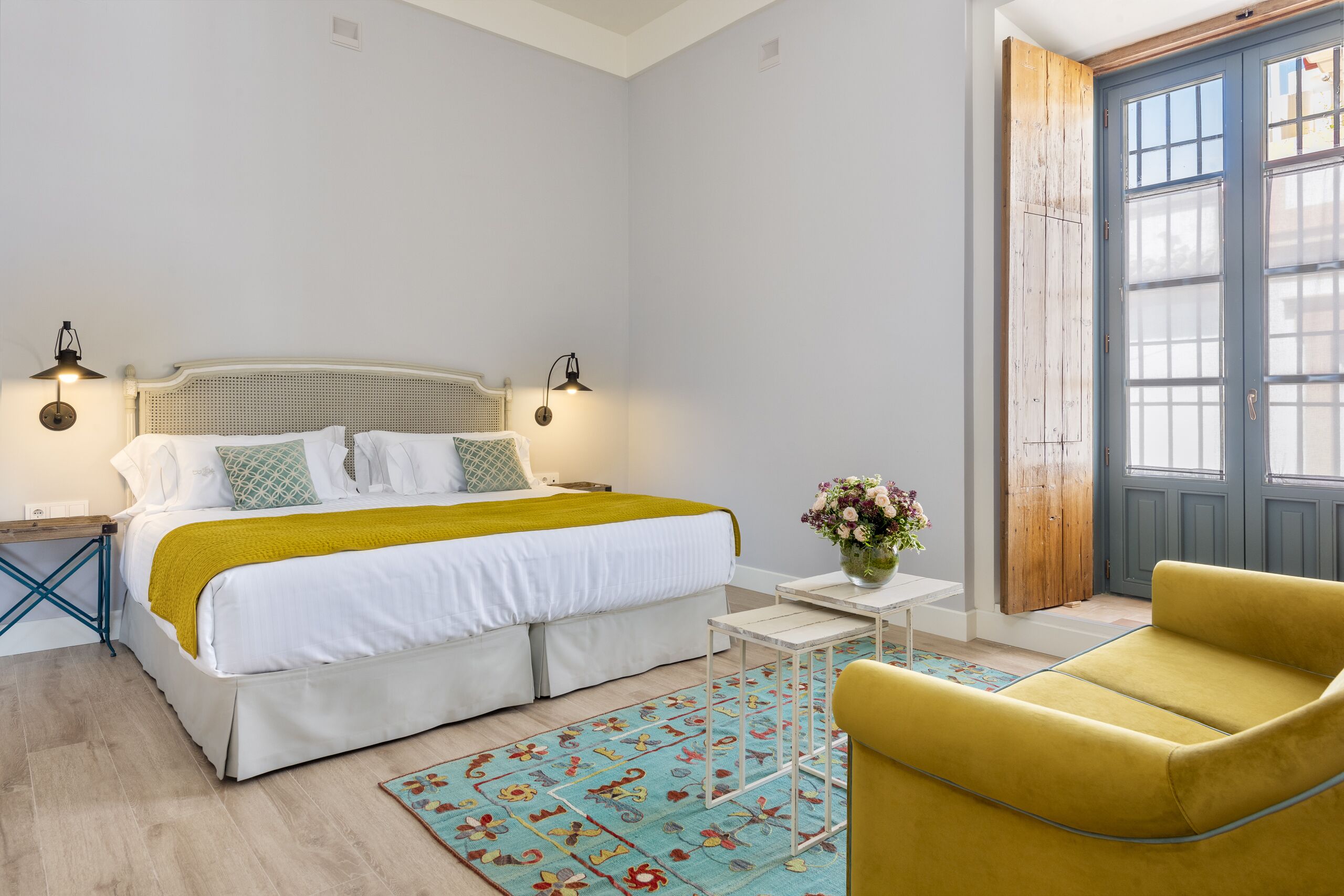 A bedroom with a bed, bedside tables, a yellow armchair, a small table with a vase, and a wooden door.
