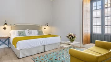 Un dormitorio con una cama, mesitas de noche, un sillón amarillo, una pequeña mesa con un florero y una puerta de madera.