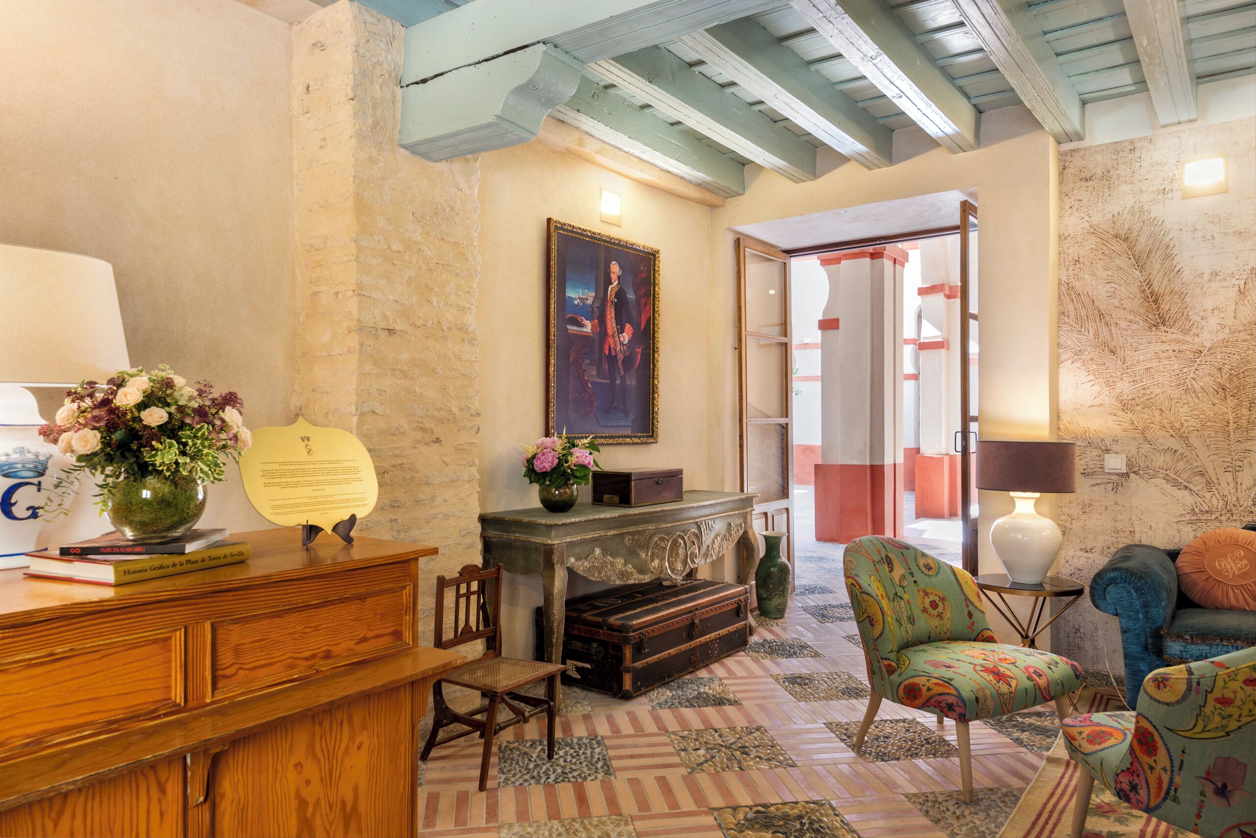 Una habitación con un escritorio de madera, una silla, una maleta antigua y un sillón colorido.