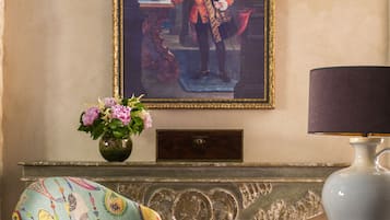 A room with a vintage chair, a framed portrait on the wall, a vase with flowers, and a lamp.