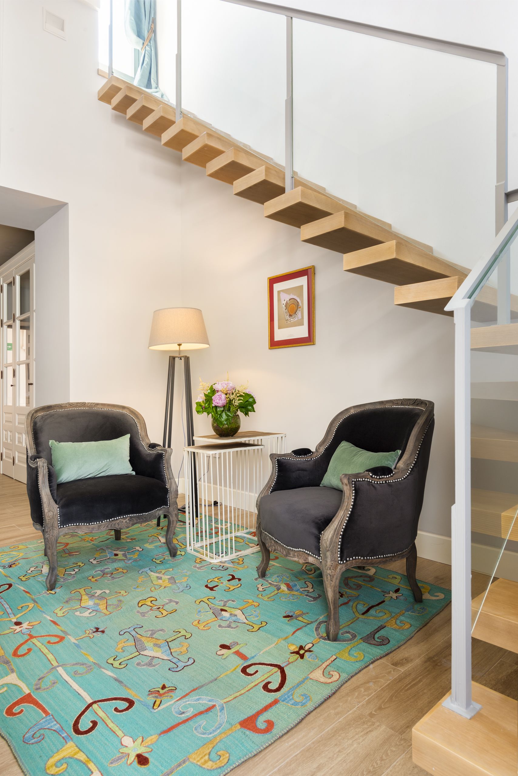 Una escalera con peldaños de madera y barandilla de vidrio, dos sillones tapizados con cojines verdes, una lámpara de pie, un cuadro enmarcado y una alfombra colorida con estampado.