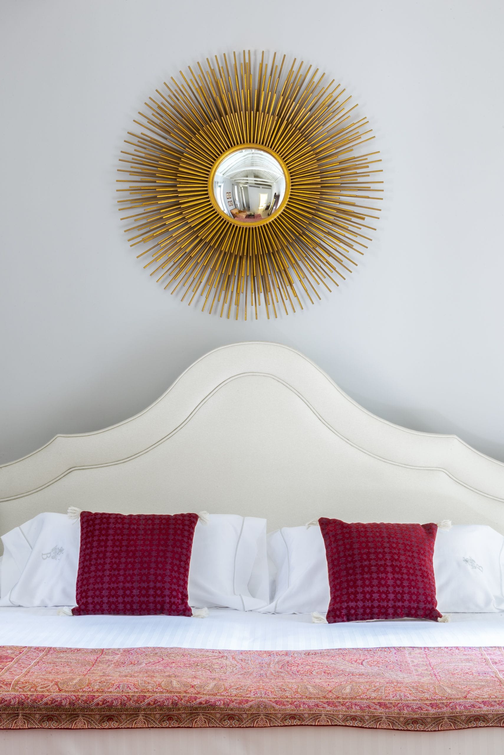 A bed with white bedding and red pillows, a white headboard, and a large gold and white sunburst mirror on the wall.