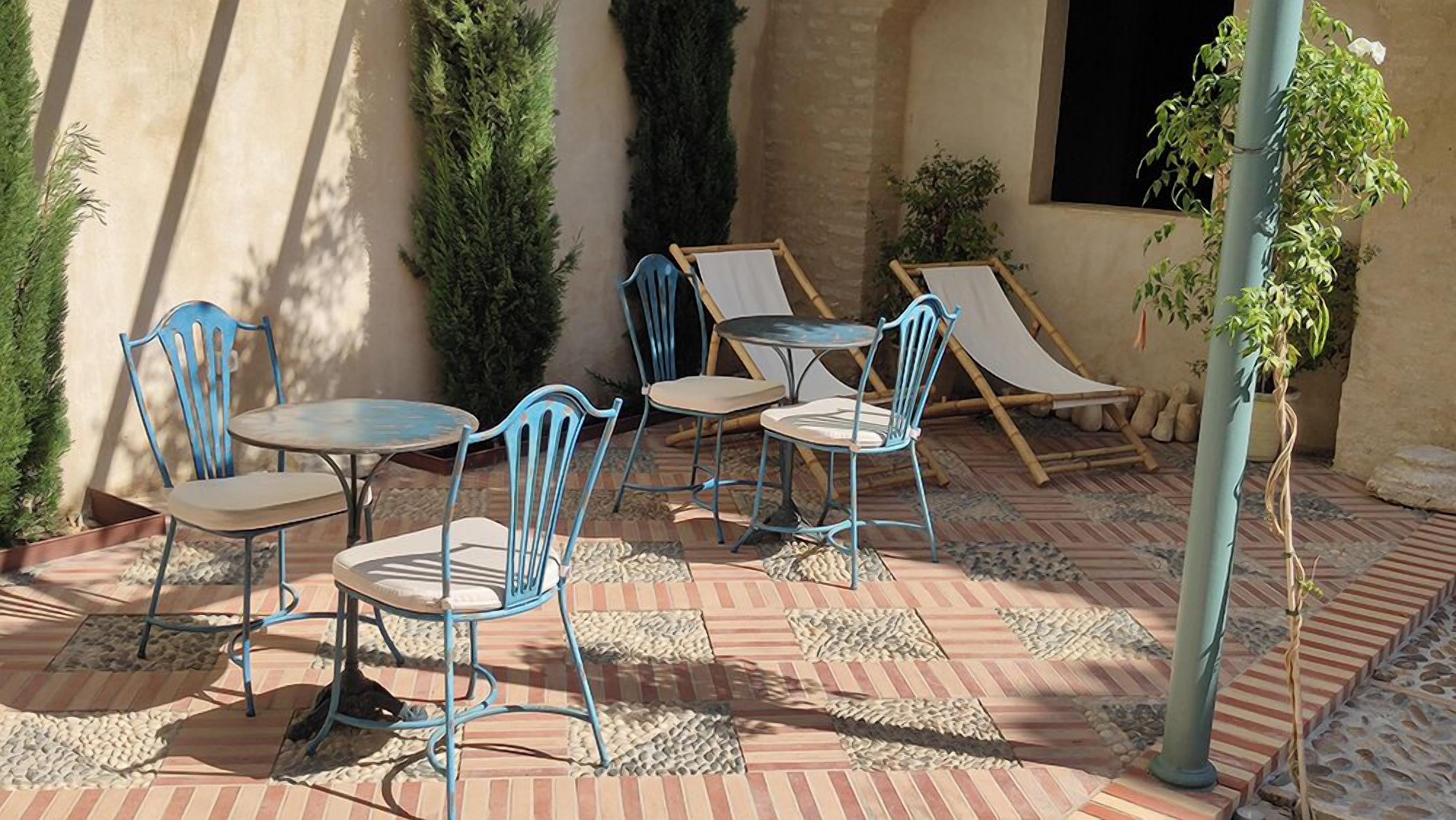 An outdoor patio with blue metal chairs and tables, surrounded by potted plants and a stone floor.