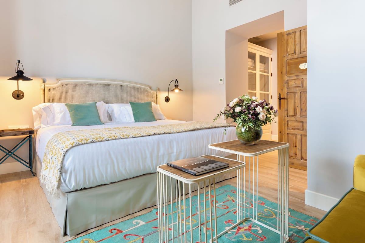 Una camera da letto ordinata con un letto, lampade sui comodini, un piccolo tavolo con un libro, un vaso con fiori e una porta in legno.