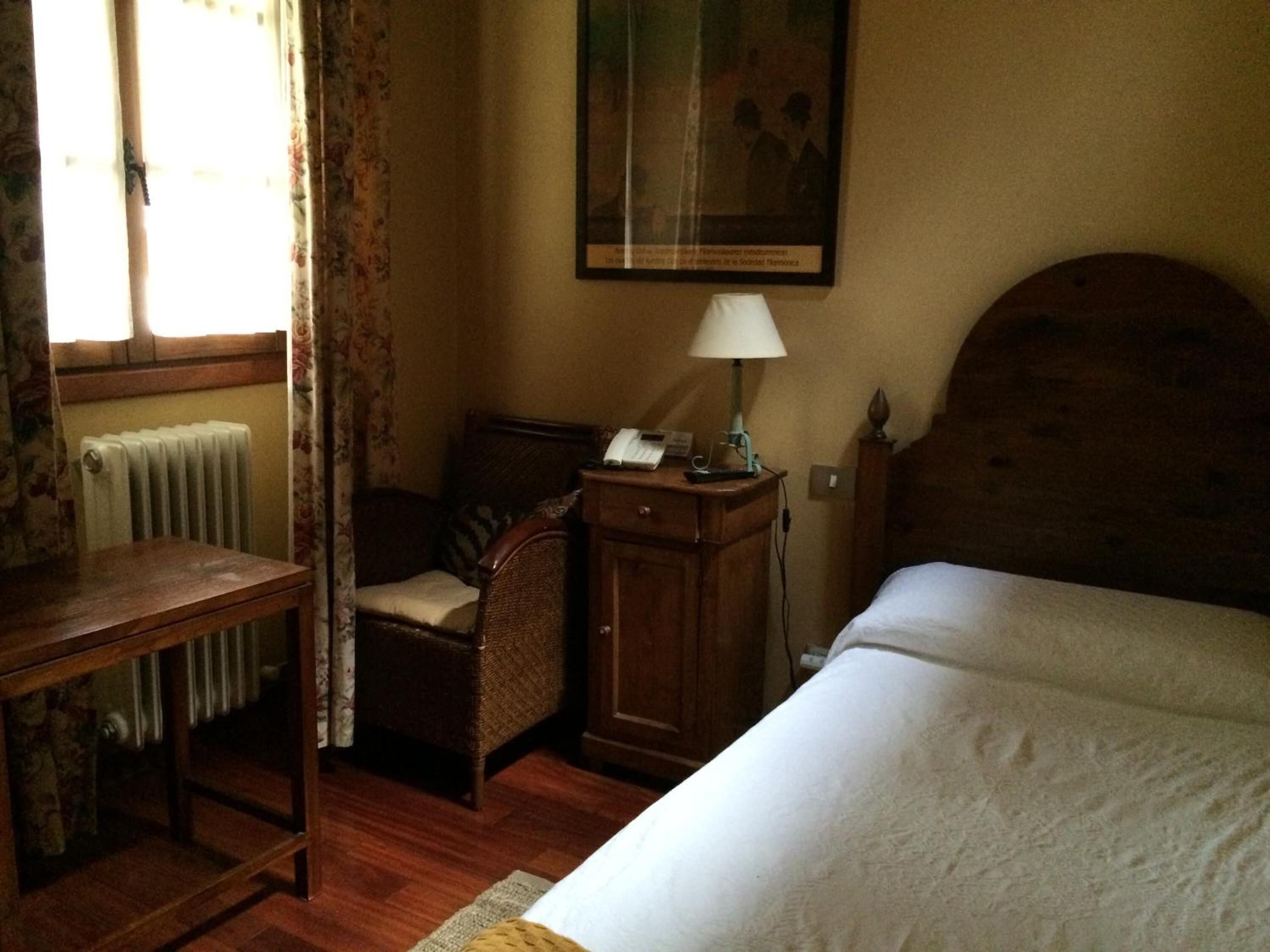 A room with a wooden bed, a small desk, a chair, a radiator, and a framed picture on the wall.