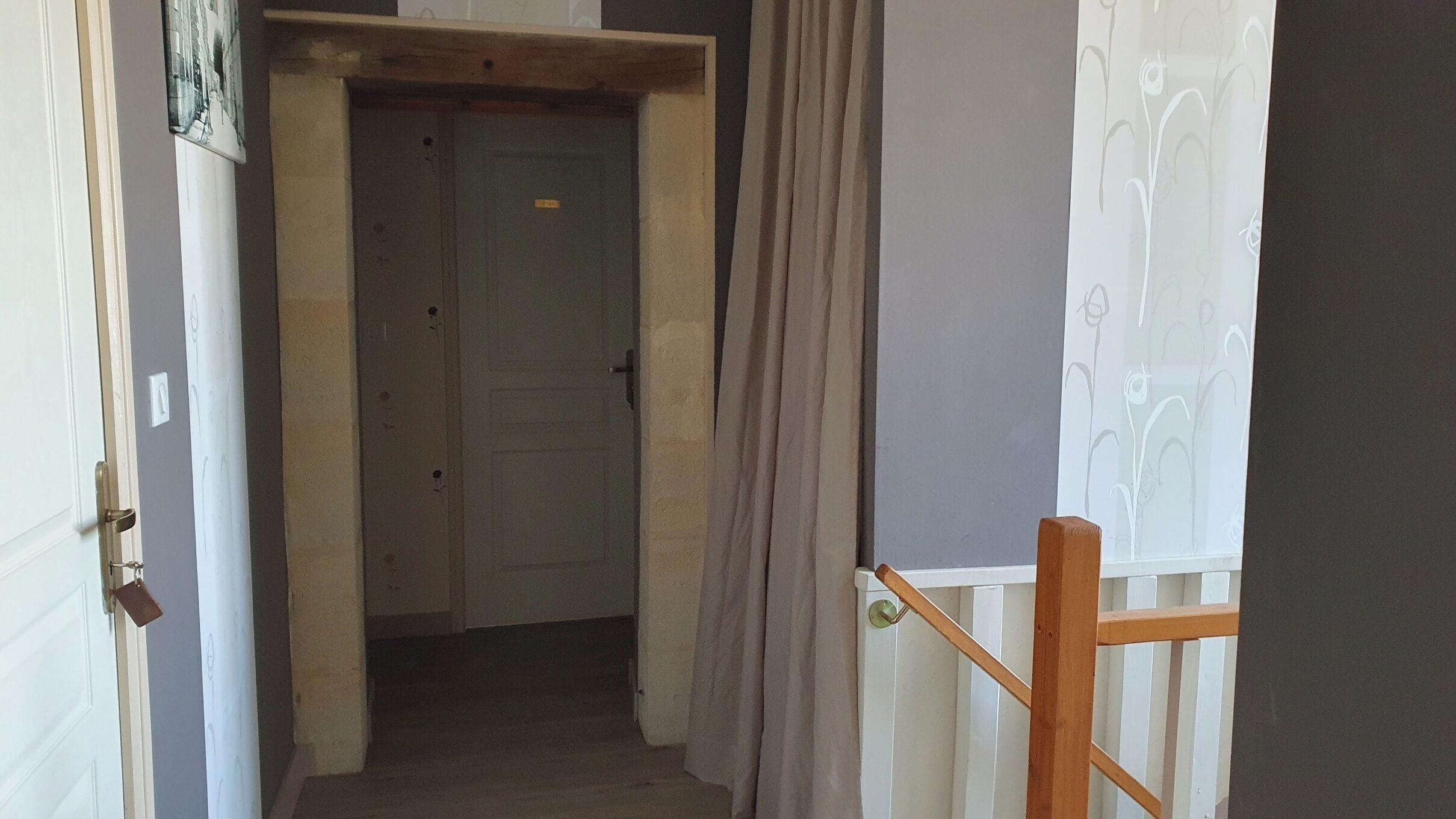 Un couloir étroit, avec une porte fermée au bout, une rambarde en bois sur le côté droit et un interrupteur sur le mur.