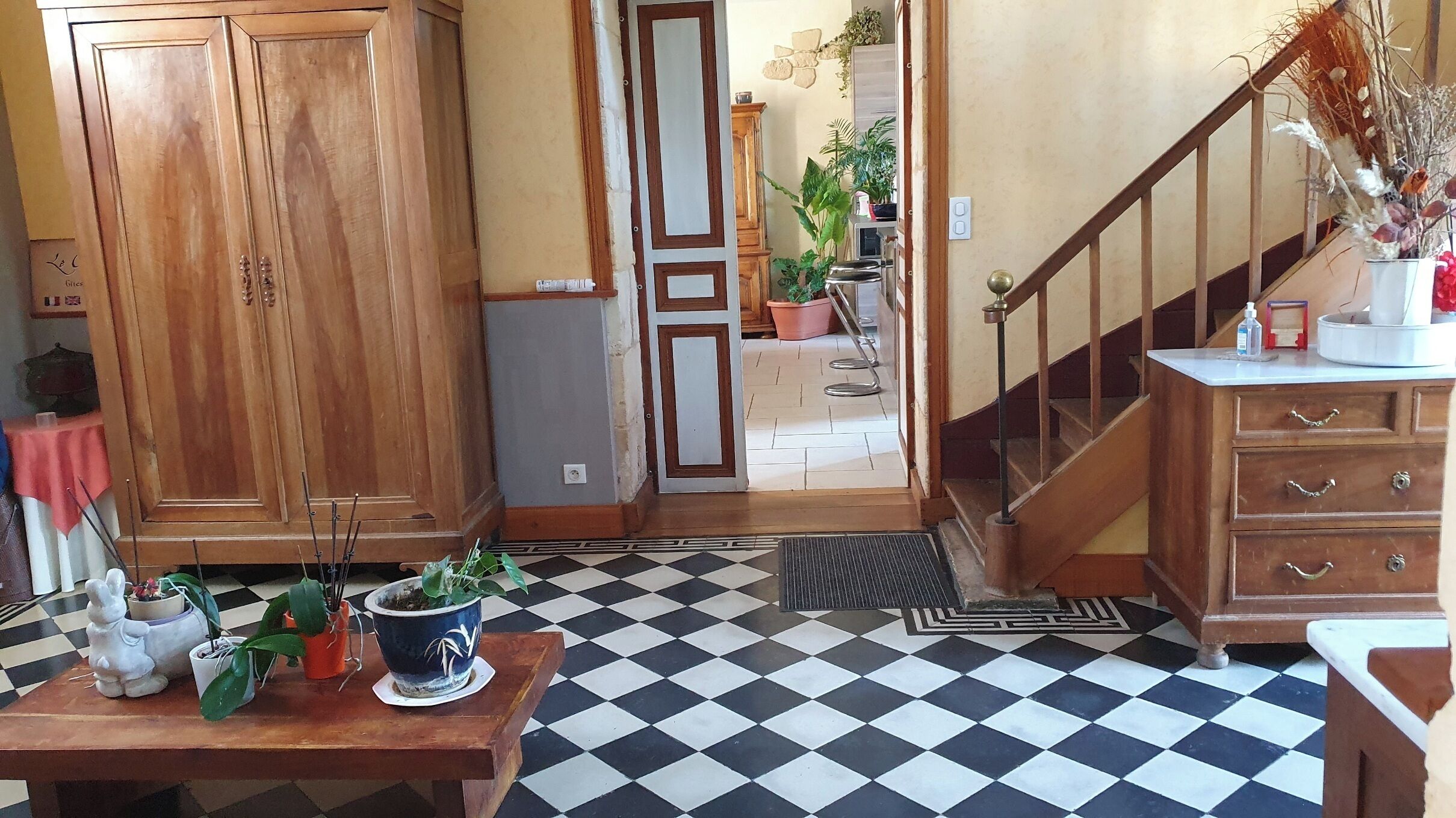 Una habitación con piso a cuadros, muebles de madera, una escalera y vista a otra habitación con plantas.