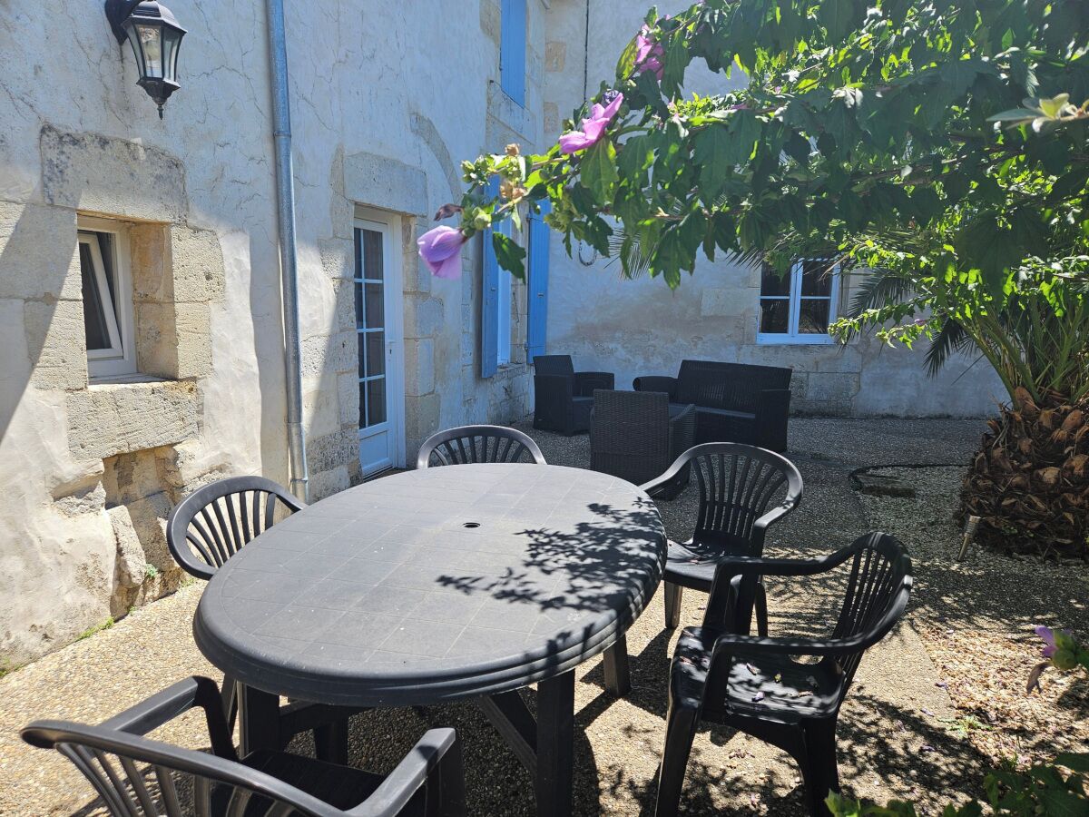 Un espace extérieur aménagé avec une table ronde et des chaises, un bâtiment en pierre doté de fenêtres, et un arbre aux fleurs violettes.
