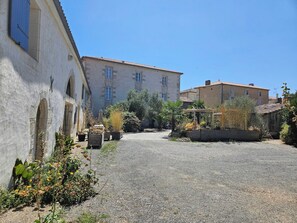 A courtyard with a gravel path, a building with arched windows, and a clear blue sky. - Chambre d'Hôtes Le Clos de Gémozac (Gémozac)