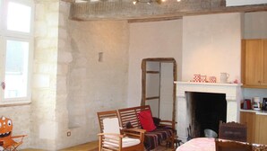 A rustic room with exposed wooden beams, a fireplace, a dining table with a patterned tablecloth, and a chair with a red cushion. - Le Manoir de la Baronnie (Boischampré)