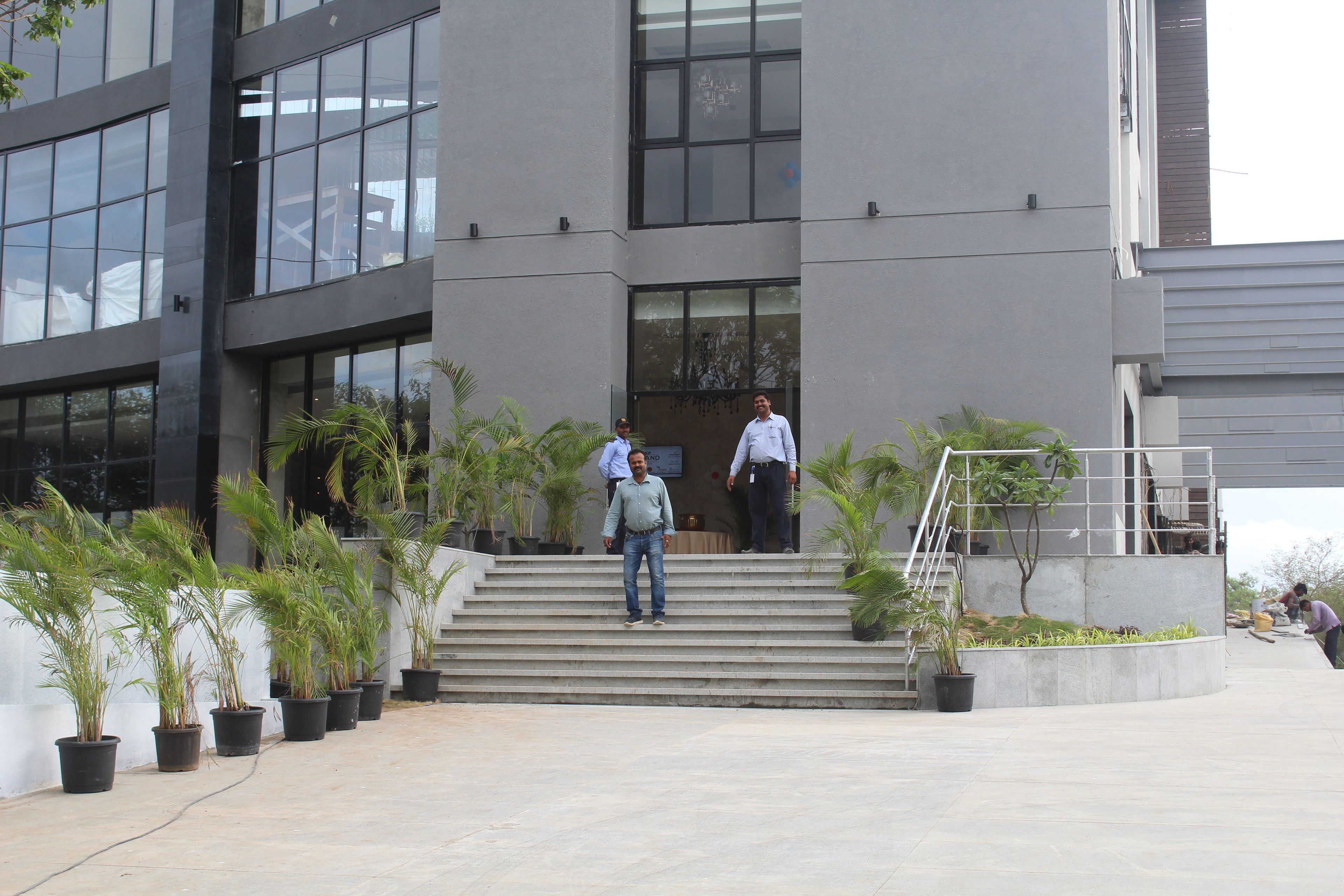 Entrada de un edificio con escalones, plantas en macetas y algunas personas.