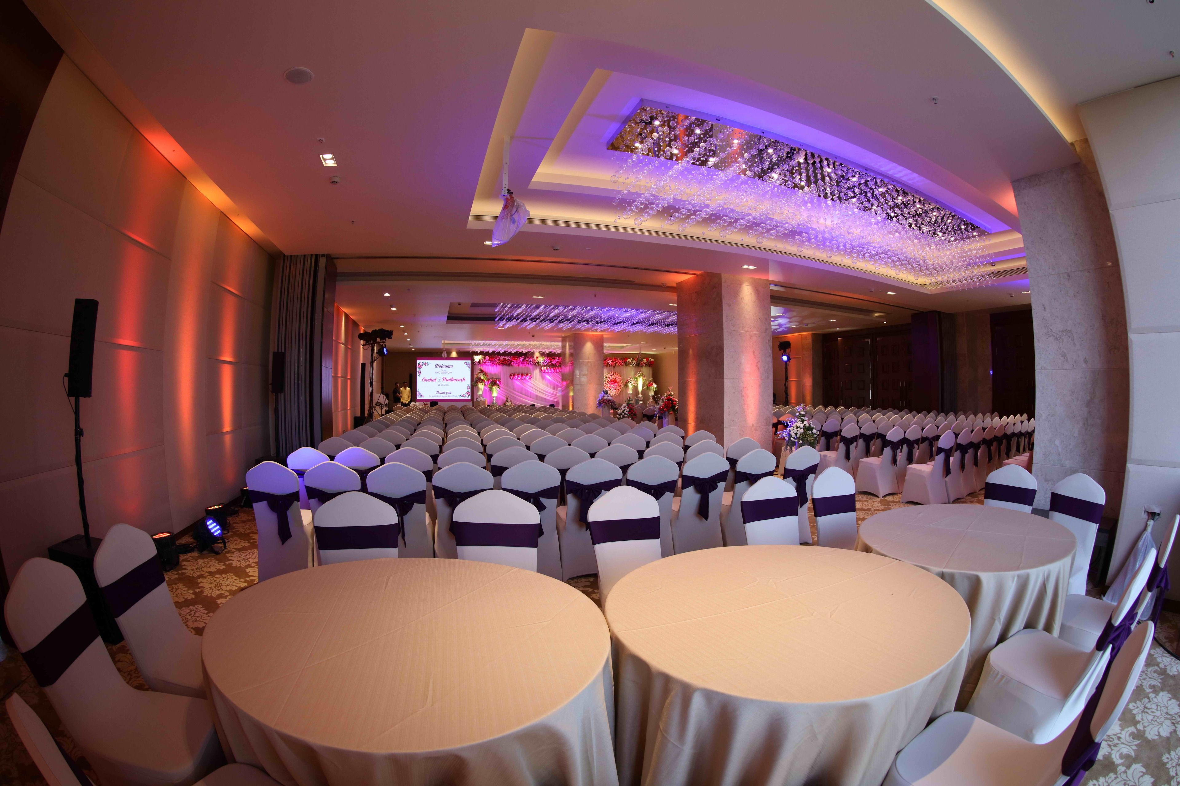 Une salle de banquet avec des rangées de tables rondes et de chaises, une scène dotée d’un écran de présentation et un éclairage décoratif.