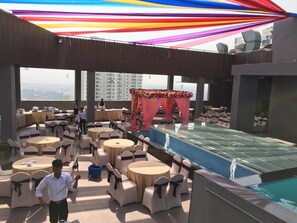 A rooftop bar with round tables and chairs, a pool, and a stage with a colorful backdrop. - Grand Tamanna Hotel Hinjawadi Pune (Paud)