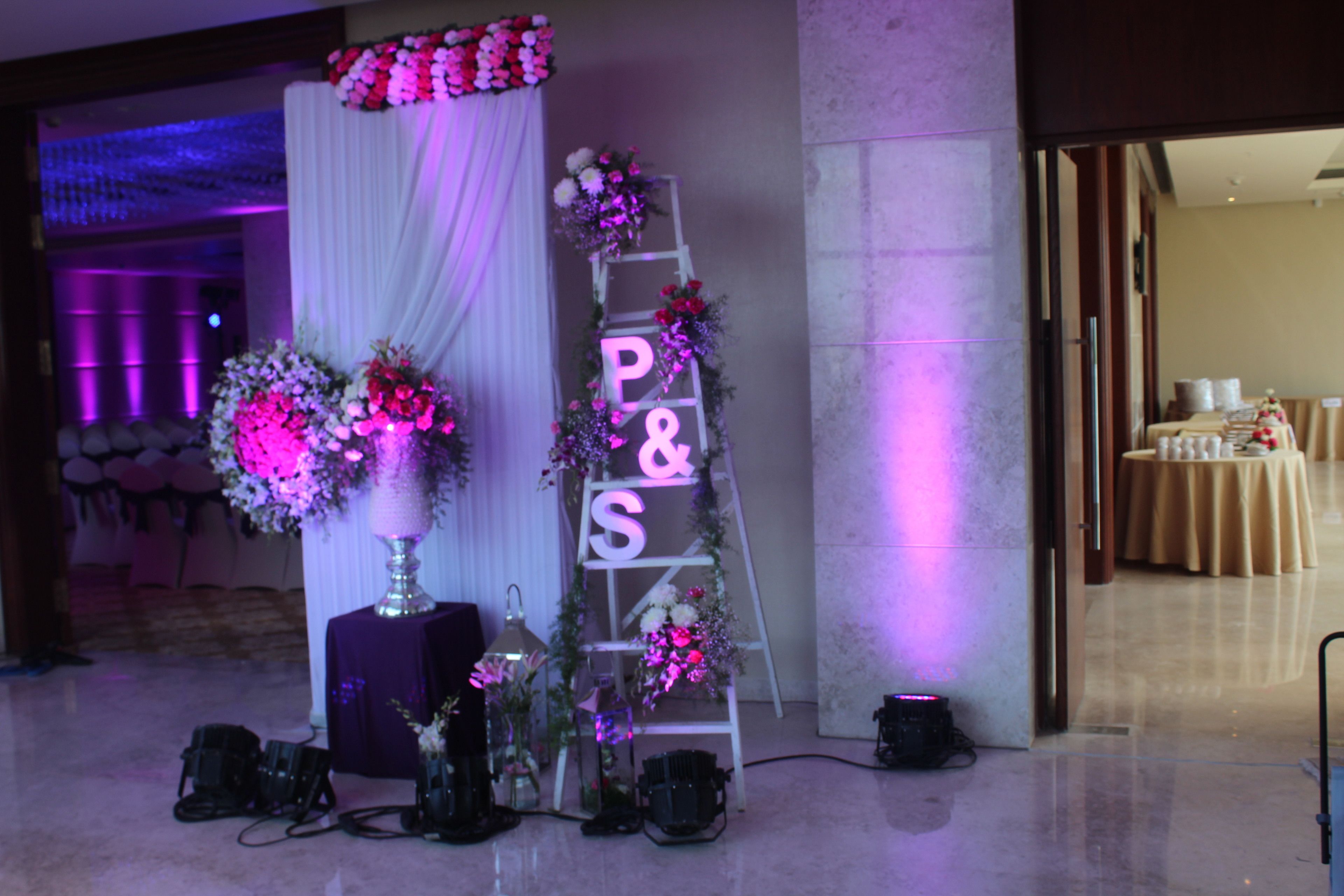 A reception area with a white draped backdrop, a floral arrangement, and a 'P&S' sign on a ladder.