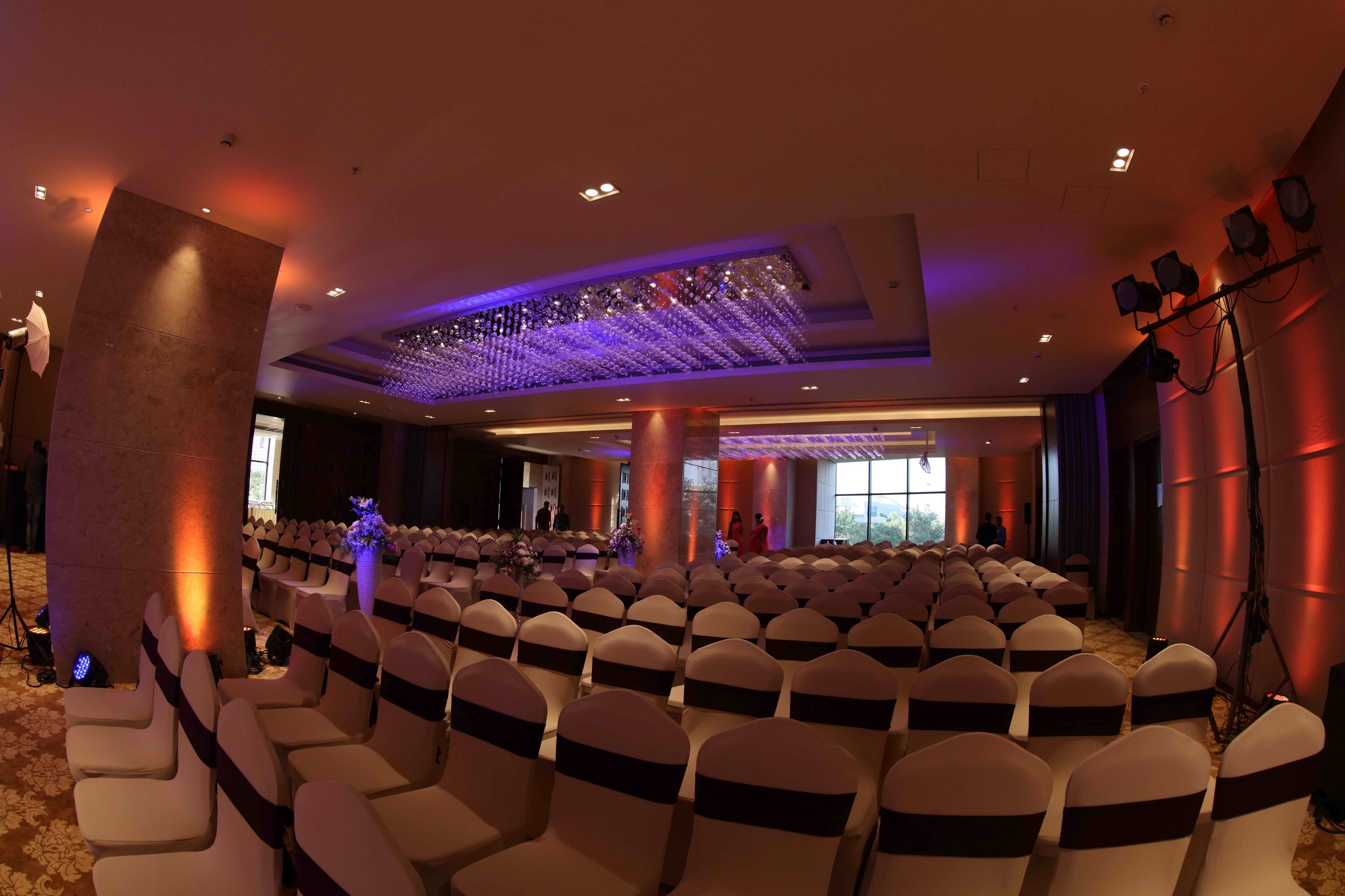 A conference room with rows of white chairs with dark sashes, a stage with a few people, and a large pillar.