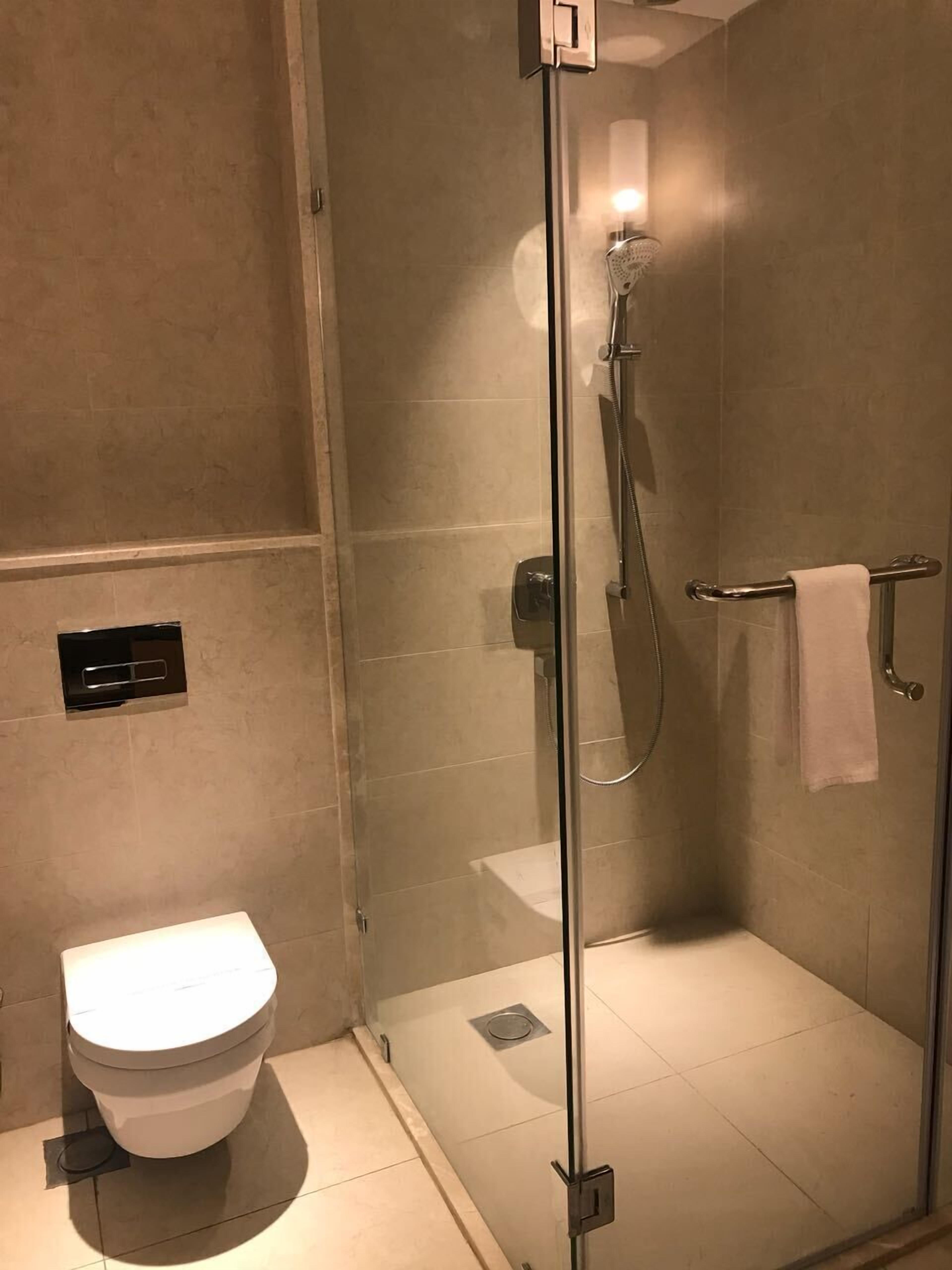 A glass-enclosed shower with a handheld showerhead, a towel rack with a towel, and a toilet with a flush button.