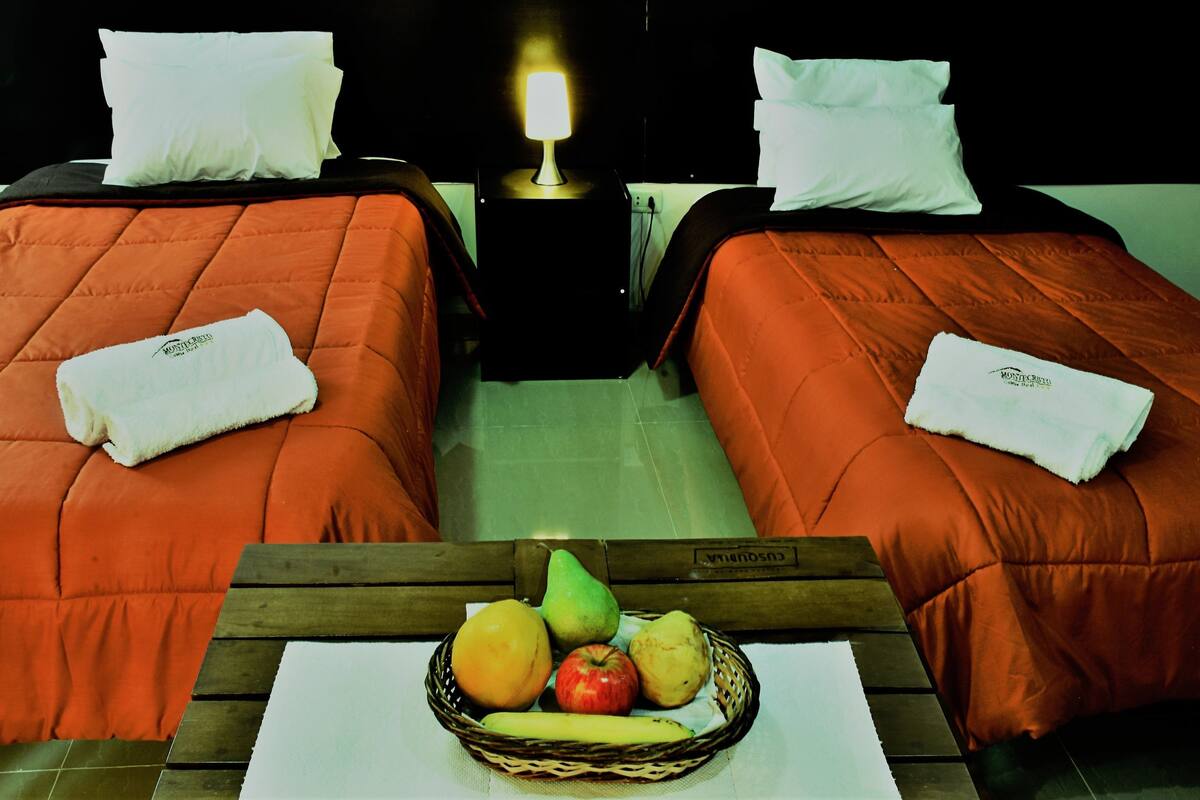 Two single beds with orange bedding, white pillows, and a bedside table with a lamp. A fruit basket with apples, pears, and a banana is on the table between the beds.