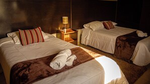 Dos camas con sábanas blancas, almohadas a rayas y una manta marrón. Hay una mesita de noche con una lámpara entre las camas.