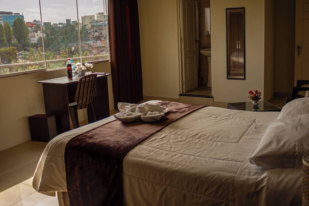 A hotel room with a large bed, a desk with a chair, a window with a view of buildings, and a bathroom visible through an open door.