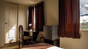 Habitación de hotel con cama, escritorio con lámpara, silla, televisor y ventana con cortinas.