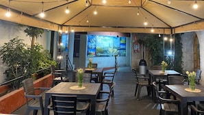 Un área de comedor al aire libre con mesas y sillas, rodeada de plantas y un mural grande al fondo.
