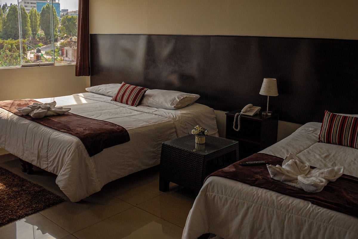 A hotel room with two beds, a nightstand, a small table, and a window with a view of buildings and trees.