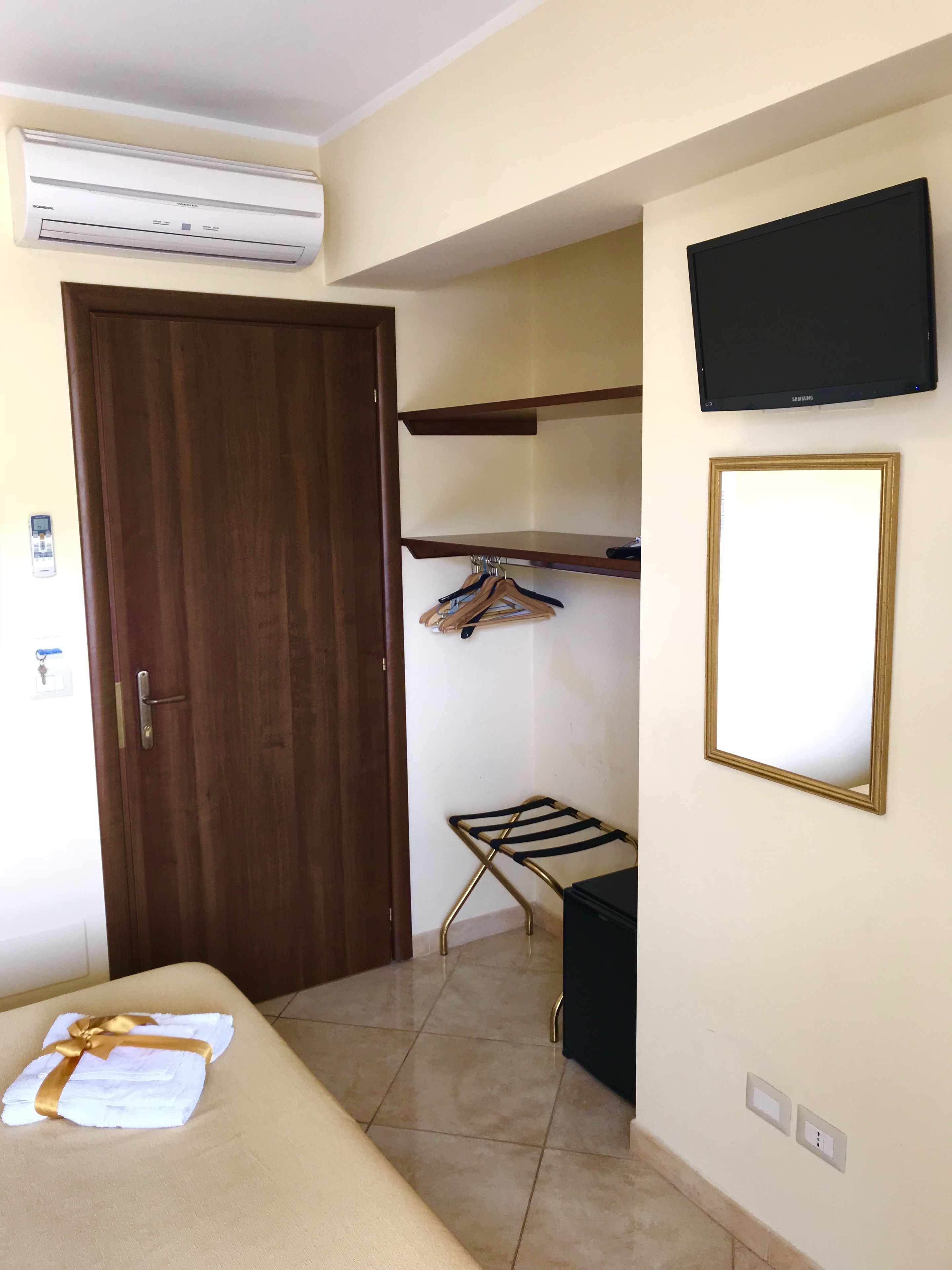 A hotel room with a bed, an air conditioning unit, a TV, a wooden door, a coat rack, and a small table.