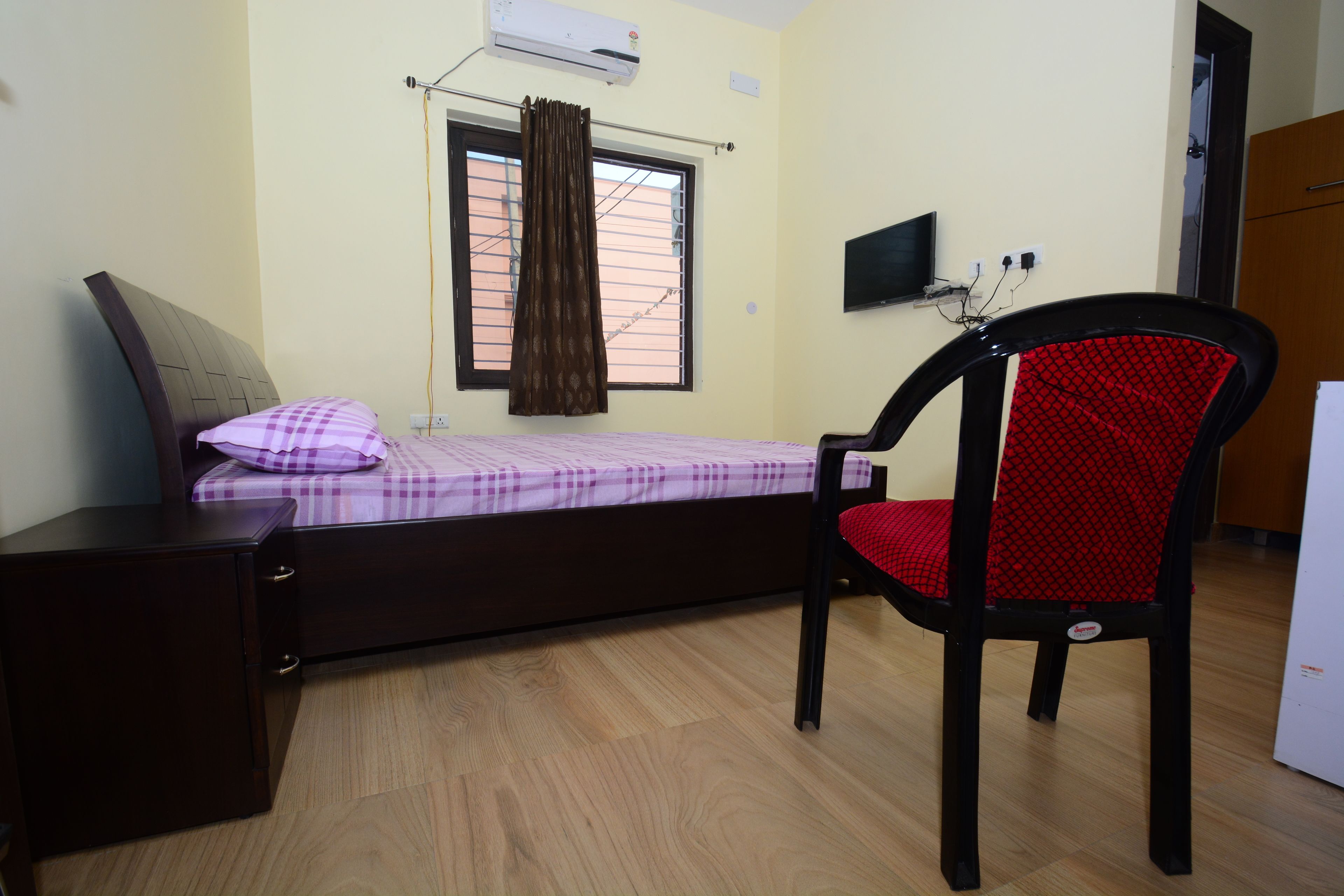 A bedroom with a bed, a desk, a chair, an air conditioner, a window with curtains, and a television on the wall.