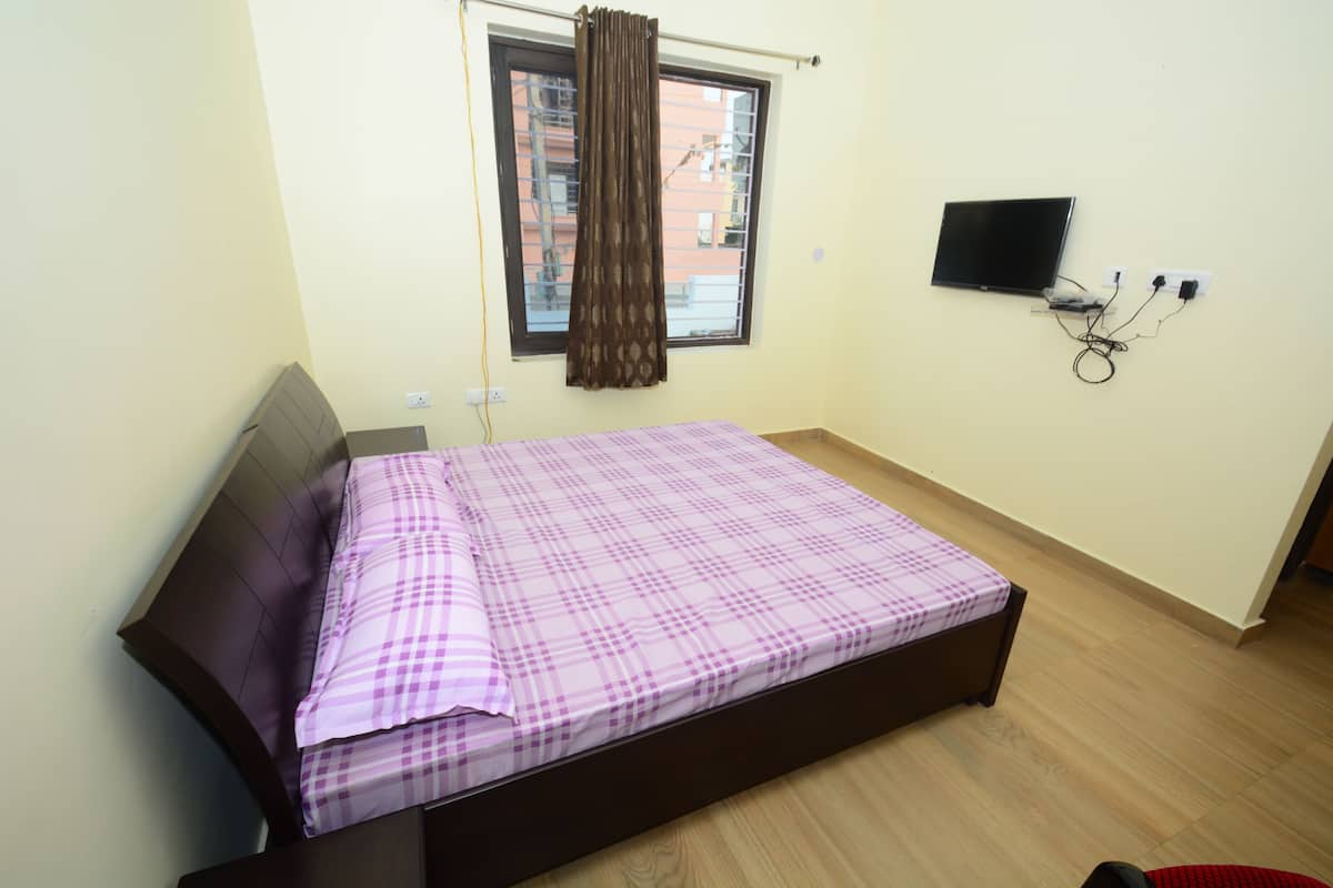 A single bed with a checkered bedspread, a window with a curtain, a wall-mounted TV, and a power outlet with cables.