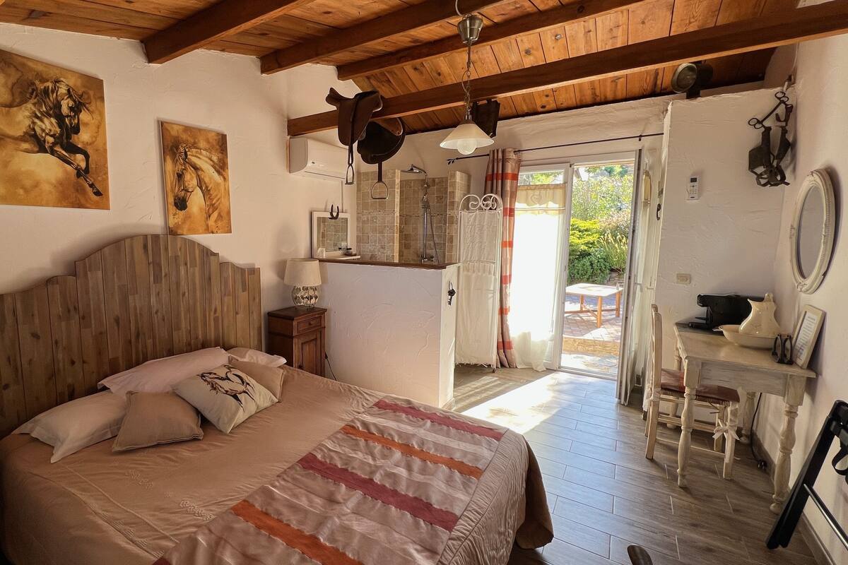 Une chambre à coucher avec une tête de lit en bois, un lit recouvert d’une couverture à rayures, une table de chevet avec une lampe, et une salle de bain visible à travers une porte ouverte.