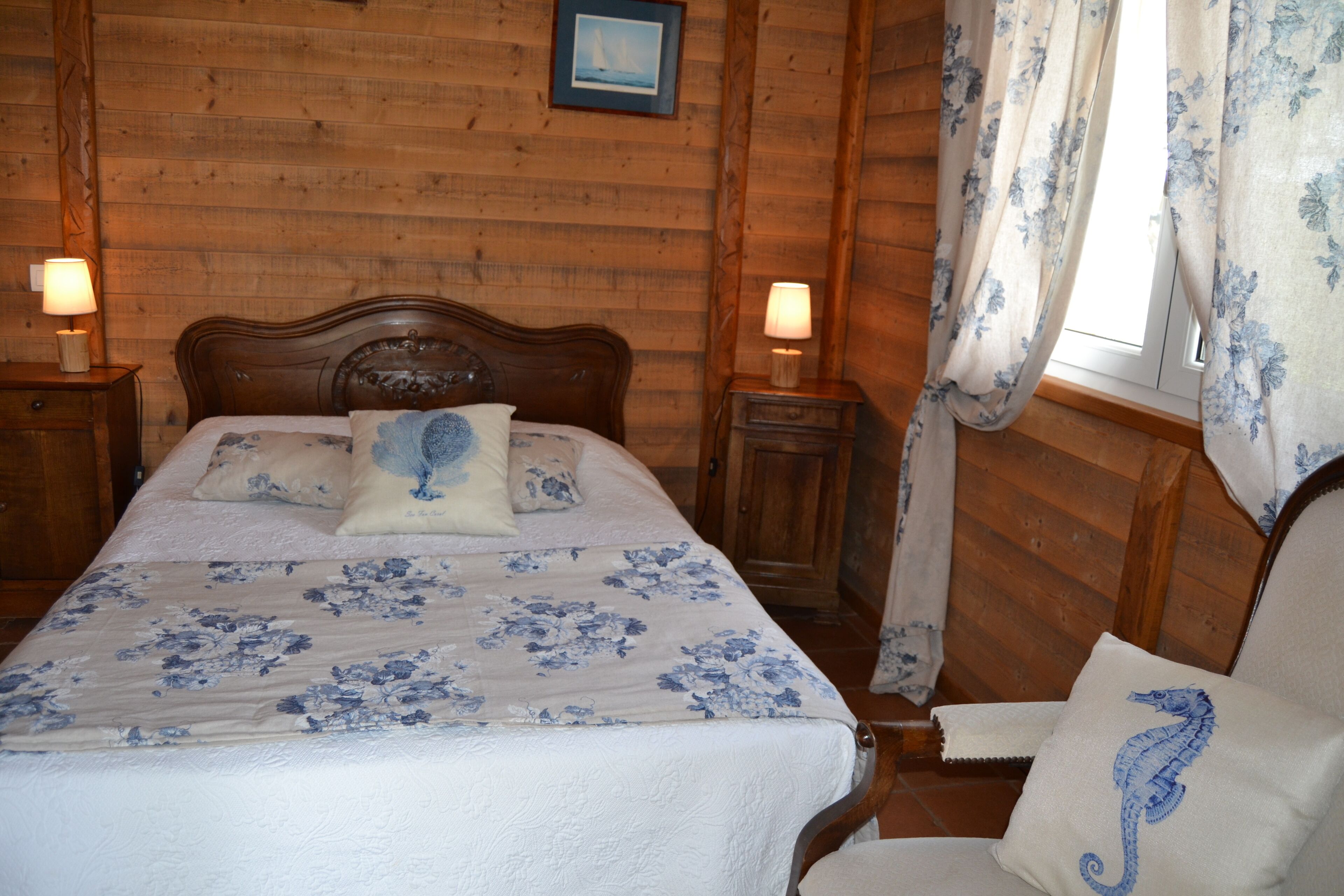 A wooden-paneled room with a bed, two bedside tables with lamps, a window with floral curtains, and a chair with a seahorse pillow.
