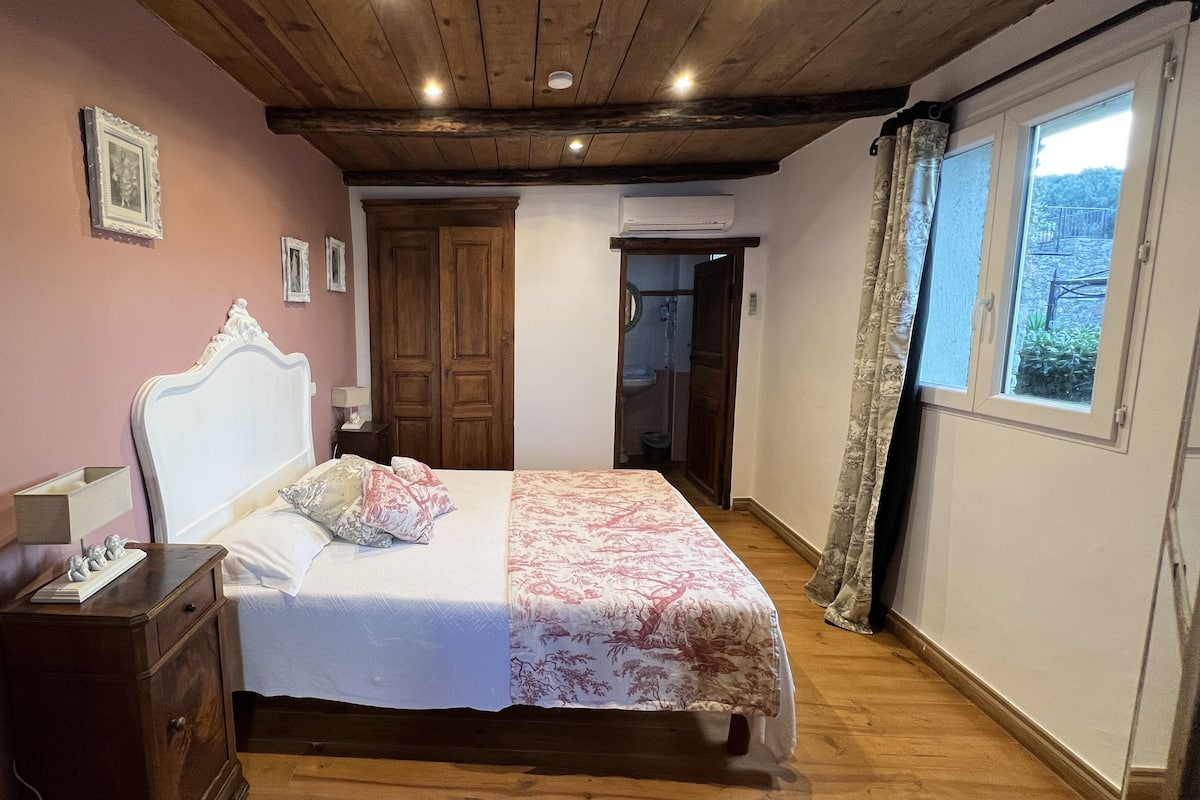 Une chambre avec un plafond en bois, un lit recouvert d’une couvre-lit à motifs floraux, une table de chevet, une commode et une fenêtre avec des rideaux.