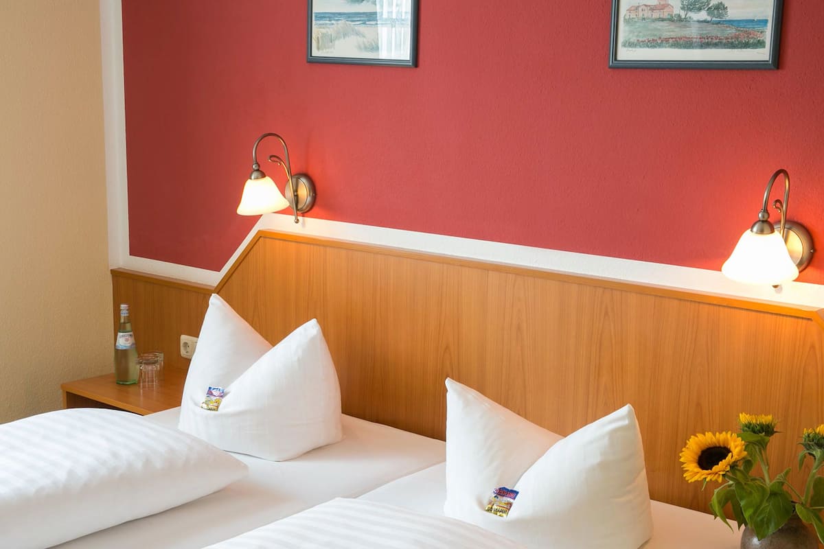 A hotel room with two beds, white bedding, a wooden headboard, two wall-mounted lamps, a nightstand with a bottle and glasses, and a vase with sunflowers.