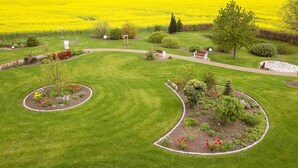 A well-maintained garden with a central lawn, surrounded by flower beds and a field of yellow flowers in the background. - Hotel Pommernland (Anklam)