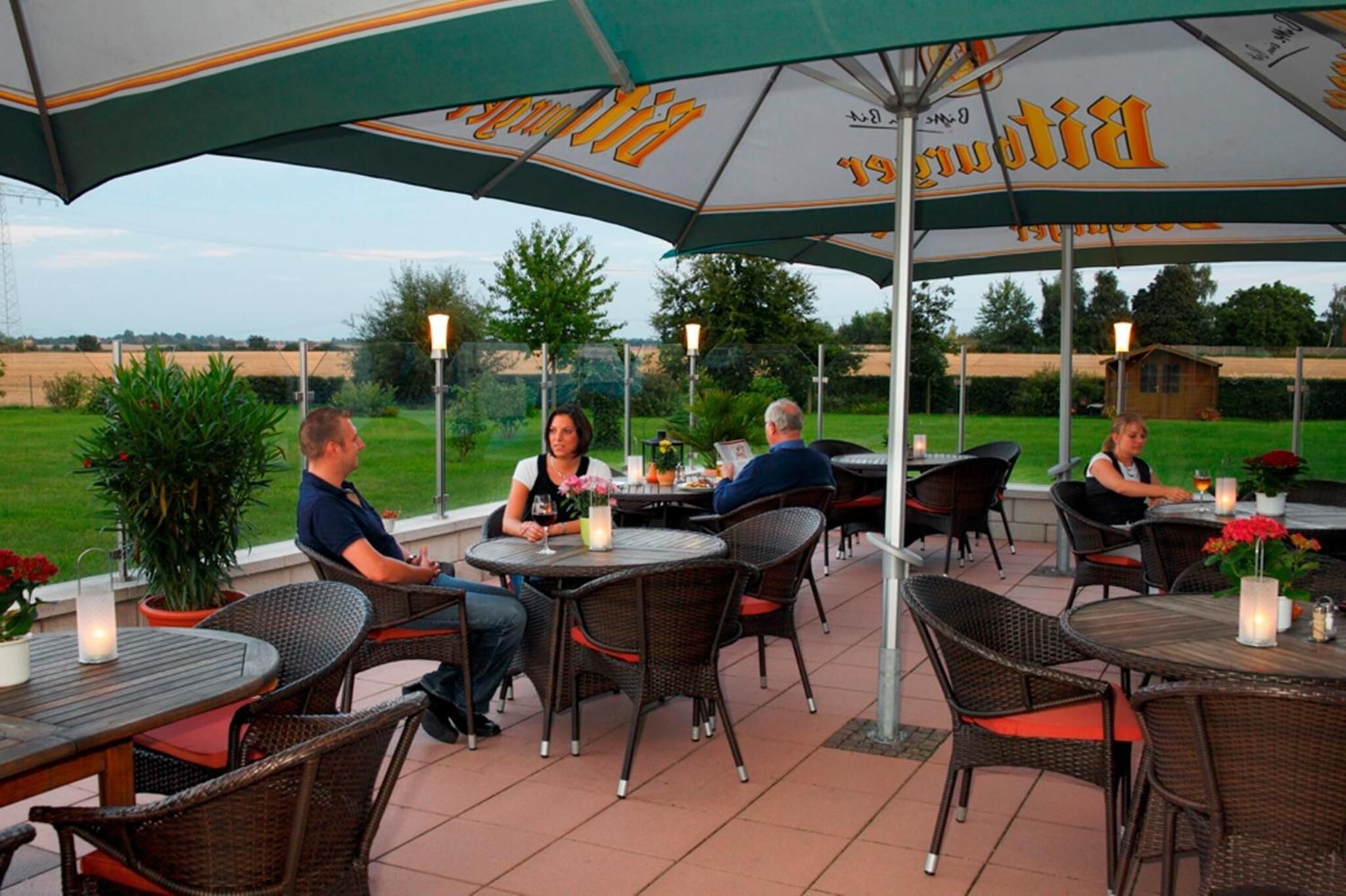 Een terras buiten met ronde tafels en rieten stoelen onder parasols, met uitzicht op het landschap.
