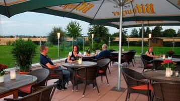 Een terras buiten met ronde tafels en rieten stoelen onder parasols, met uitzicht op het landschap.