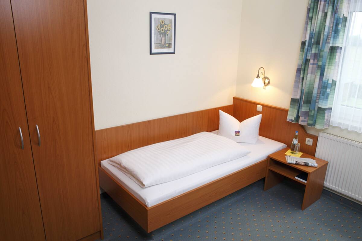 A single bed with a wooden headboard and footboard, a bedside table with a lamp, a small shelf, and a framed picture on the wall.