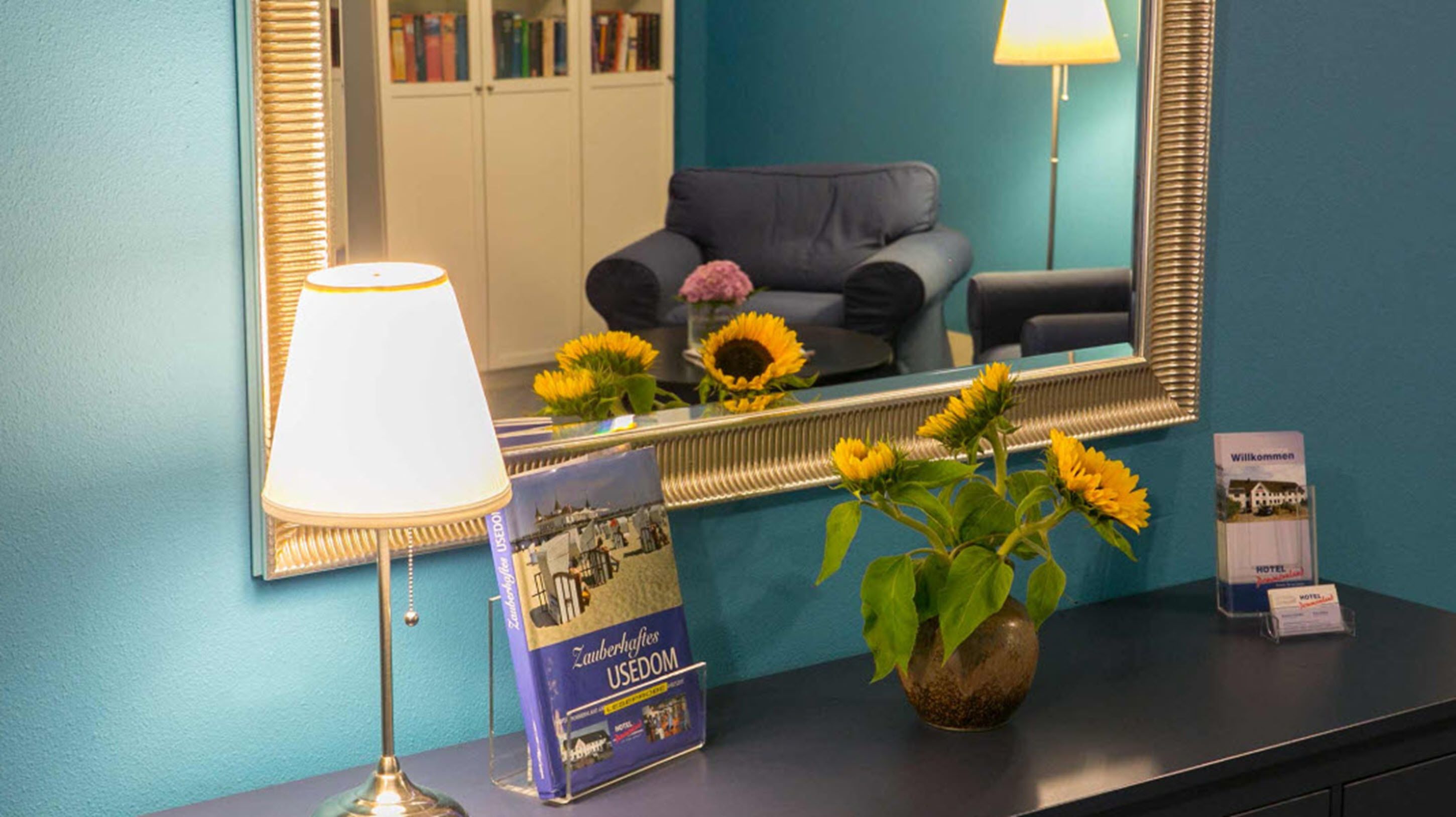 A reception desk with a lamp, a welcome sign, a brochure, and a vase with sunflowers.