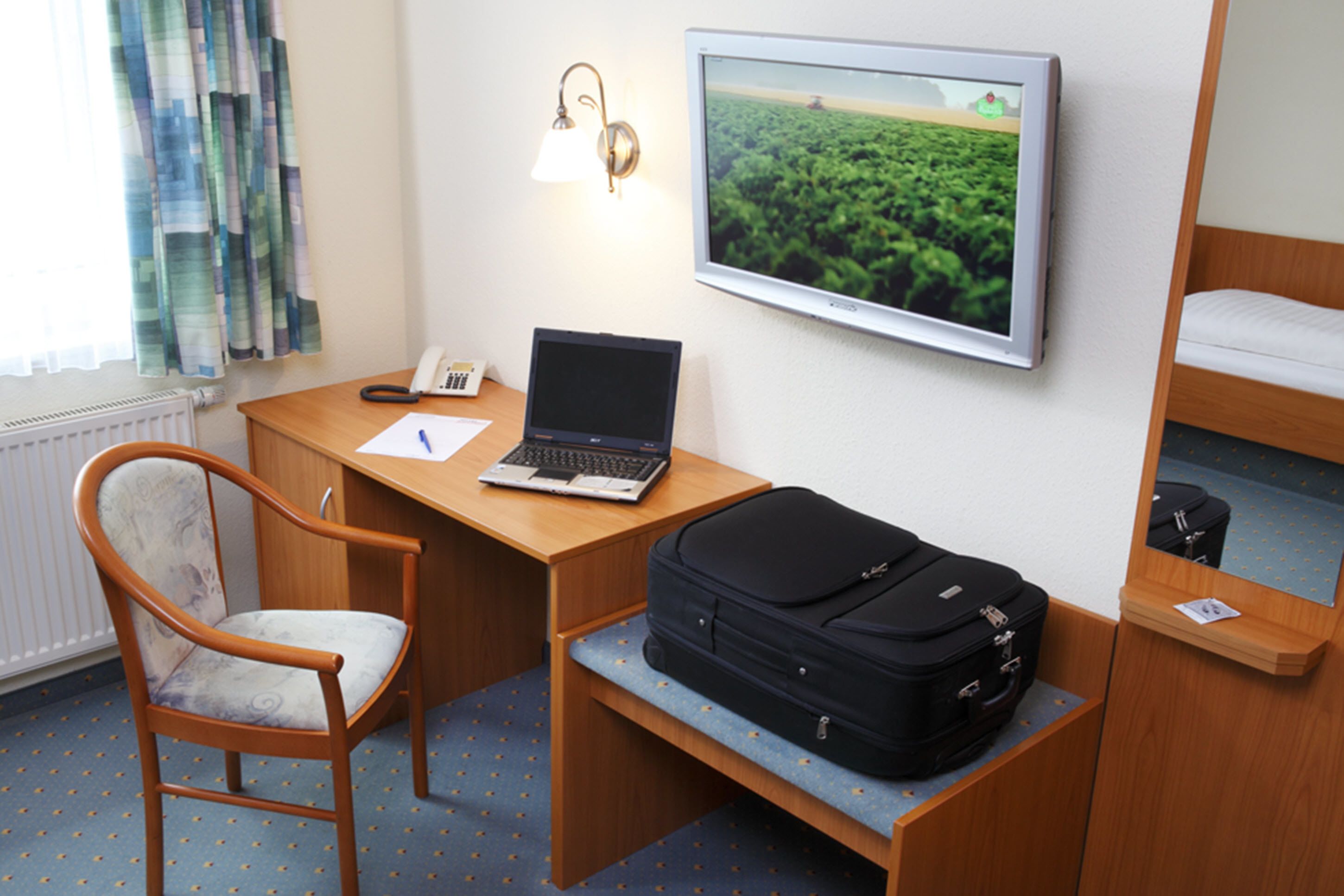 A hotel room with a wooden desk, a laptop, a chair, a bed, a suitcase, a wall-mounted TV, and a window with curtains.