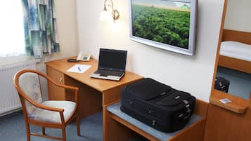 A hotel room with a wooden desk, a laptop, a chair, a bed, a suitcase, a wall-mounted TV, and a window with curtains.
