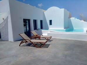Um edifício branco com piscina, duas espreguiçadeiras e uma mesa com uma cesta de frutas.