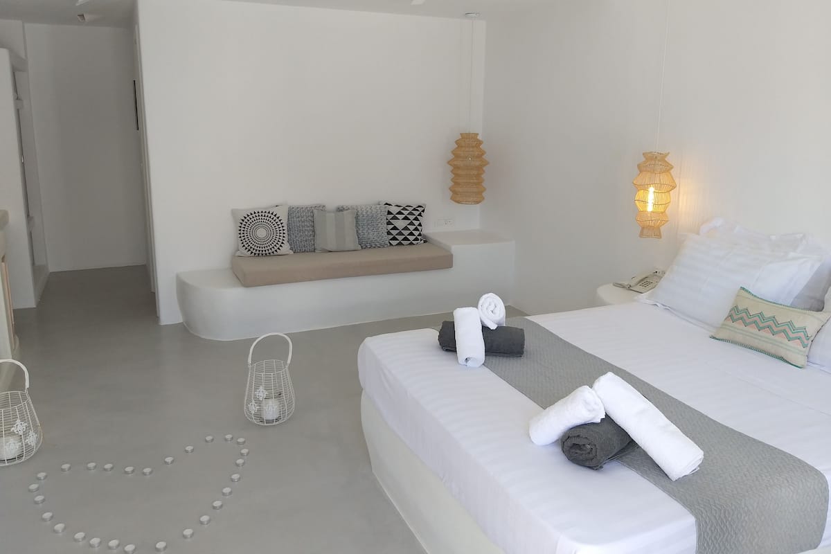 Una habitación de hotel moderna con una cama grande, una zona de estar con cojines, una mesita de noche con toallas y una lámpara de pared.
