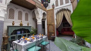 An indoor dining area with a table set for a meal, surrounded by ornate architectural details and a checkered floor pattern. - Riad Sanaa Rose (Fes)