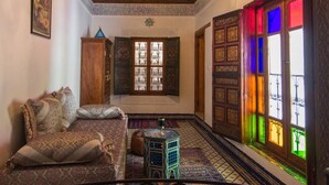 A traditional interior with a patterned ceiling, a chandelier, a sofa with cushions, a small table, and a colorful window. - Riad Sanaa Rose (Fes)
