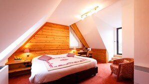 Ein Schlafzimmer mit schrägen Wänden, einem Bett mit gemustertem Überwurf, einem Schreibtisch mit Stuhl und einem Fenster.