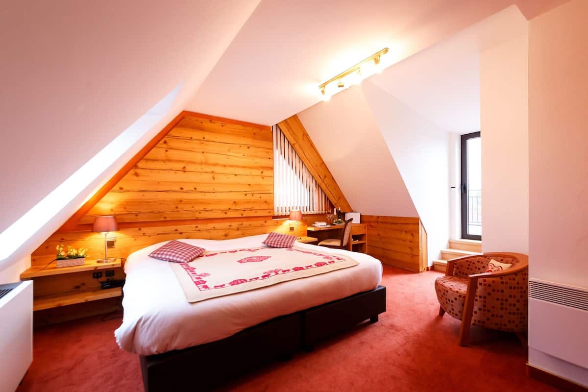 A bedroom with a sloped ceiling, a bed with a patterned bedspread, a desk with a chair, and a window.