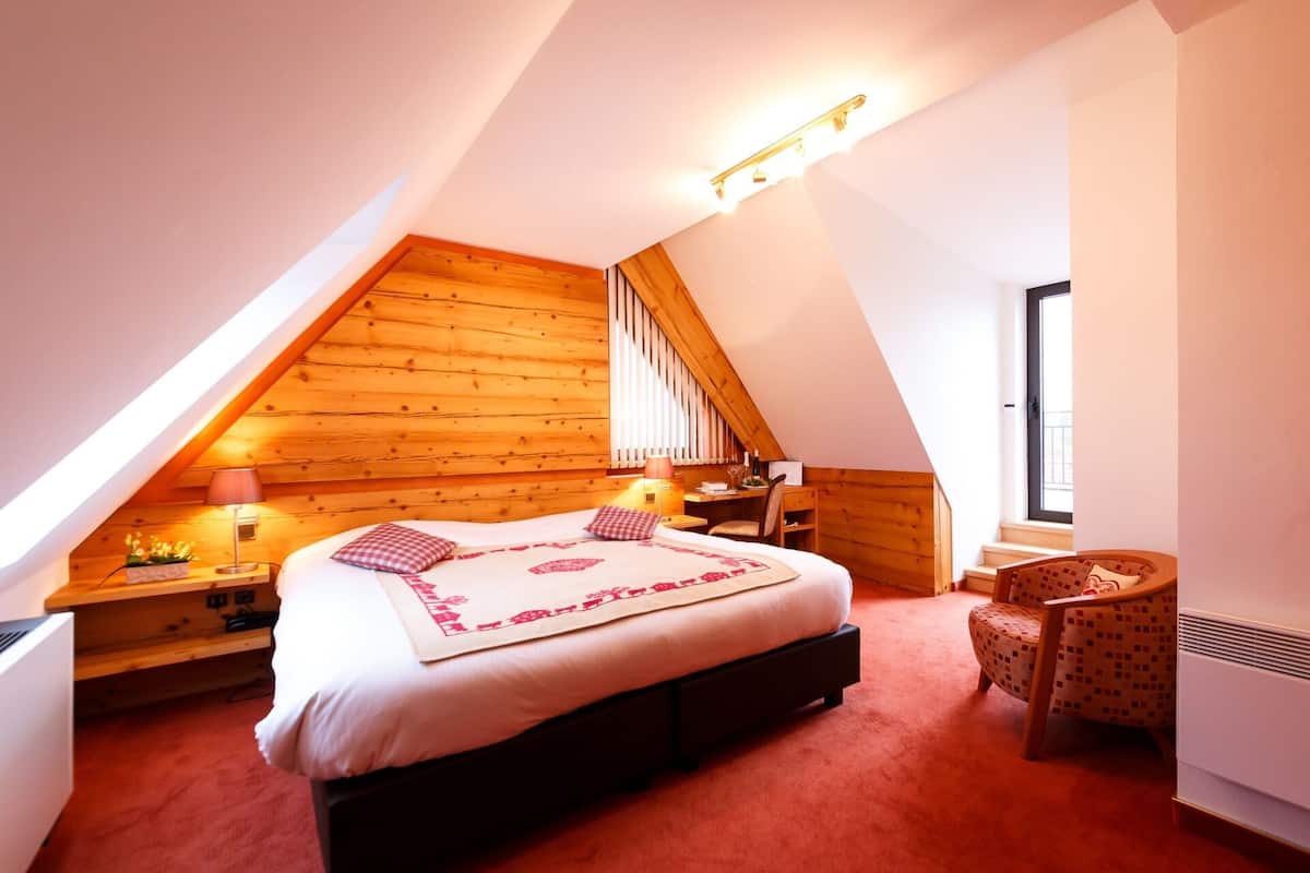 A bedroom with a sloped ceiling, a bed with a patterned bedspread, a desk with a lamp, and a chair.