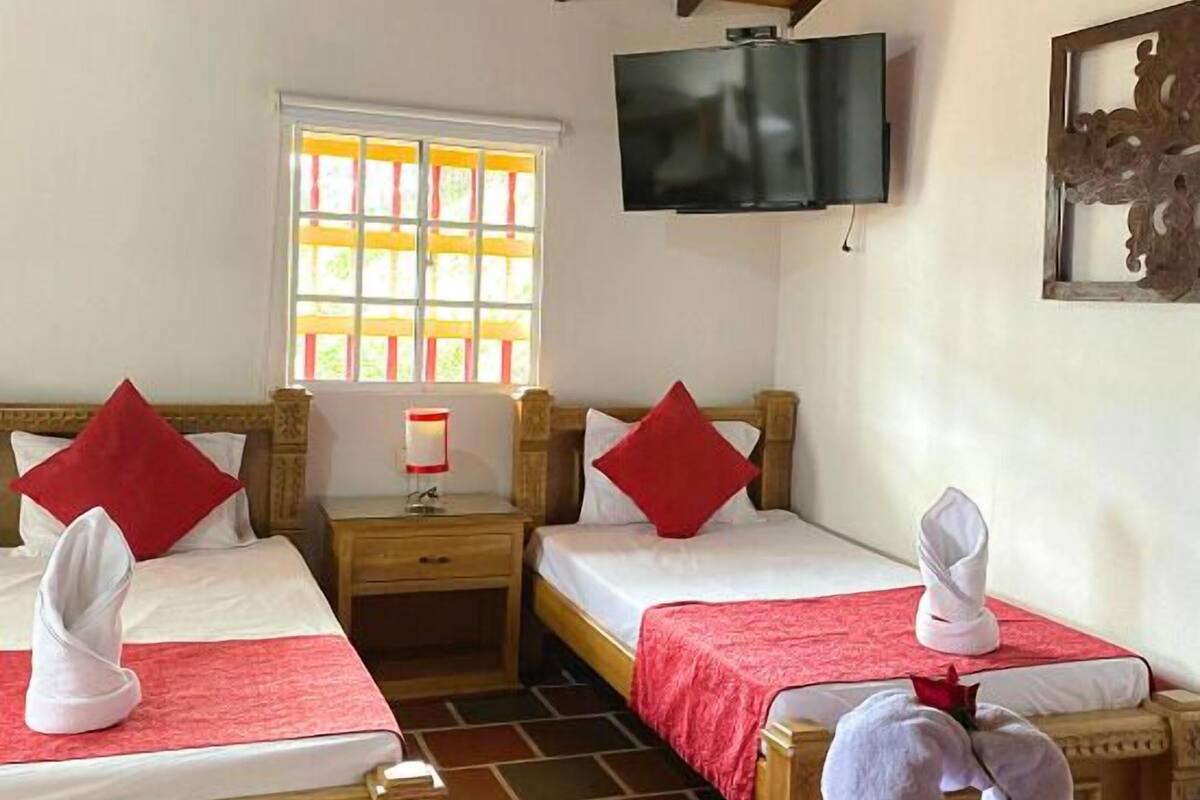 Une chambre avec deux lits, chacun recouvert d’une couverture rouge, agrémentée d’oreillers et de serviettes. Un téléviseur est fixé au mur, et une fenêtre offre une vue sur la verdure environnante.